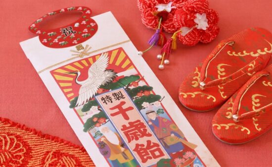 赤い背景に置かれた七五三の縁起物。鶴と亀、松竹梅が描かれた伝統的な「千歳飴」の袋と、金色の刺繍が施された赤い子供用の草履、可愛らしい花の髪飾りが並ぶ、日本のお祝いを感じさせる華やかな一枚